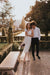 A couple stands closely in a sunlit garden. The woman, wearing the minimal silk organza Lia Dress by Noémiah, holds a bouquet, while the man in a white shirt and black pants leans affectionately on her shoulder. They're surrounded by greenery and trees.