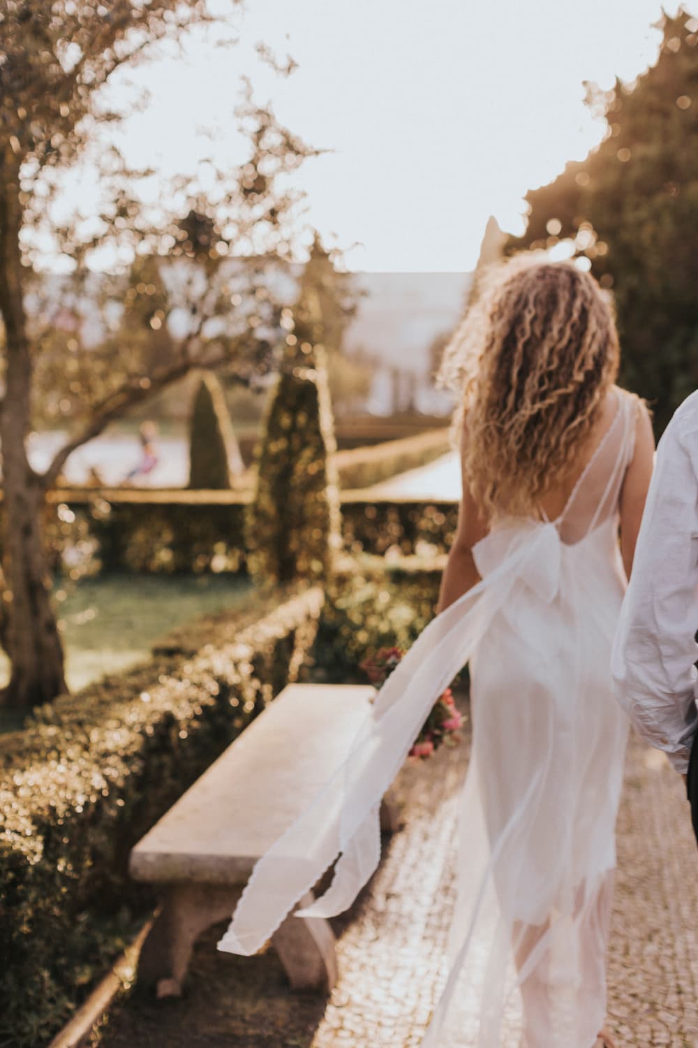 A woman with curly hair wearing the Lia Dress by Noémiah walks along a garden path, enjoying the warm sunlight. The non-traditional bride is closely followed by another person. The lush garden, framed by manicured hedges and trees, provides a serene backdrop to their journey.