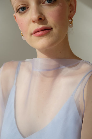 A light-skinned woman with short, blonde hair is wearing hoop earrings and a sheer top over a sleeveless pastel colored Lia Dress by Noémiah, reminiscent of a minimalist wedding dress. Her subtle smile and the soft lighting highlight her natural features, ideal for non-traditional brides.