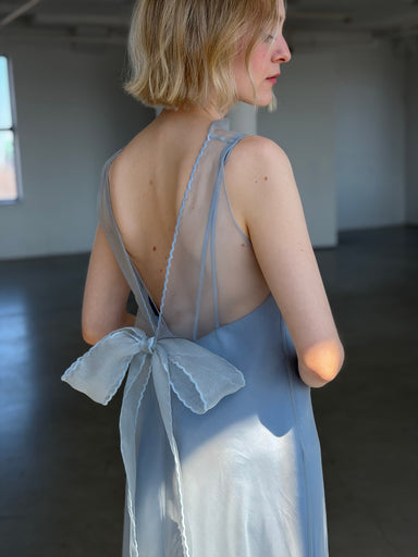 A woman with short blonde hair wears the Noémiah Lia Dress—a sheer, light blue slip dress with a low back and large bow—while standing in a sunlit, empty room with concrete floors and white walls.