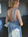 A woman with short blonde hair wears the Noémiah Lia Dress—a sheer, light blue slip dress with a low back and large bow—while standing in a sunlit, empty room with concrete floors and white walls.