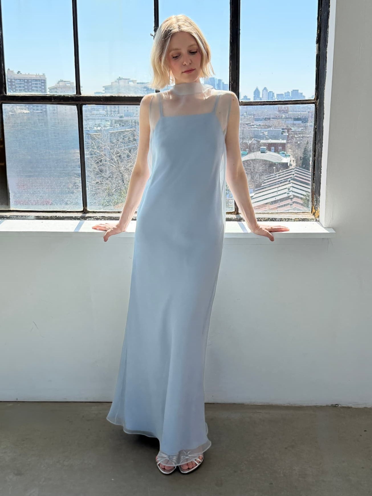 A woman wearing the Lia Dress by Noémiah stands by a large window overlooking city buildings and a clear sky, looking down and to the side with her hands resting on the windowsill.