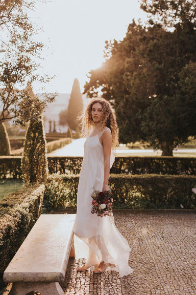 Wearing the exquisite Lia Dress by Noémiah, a woman gracefully holds a bouquet of flowers as she stands on a stone path in a sunlit garden. The setting, adorned with tall hedges and trees that cast a warm, golden glow, provides an ideal backdrop for non-traditional brides.