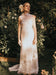A woman stands in a garden, illuminated by sunlight and surrounded by green plants and pale flowers, wearing the Lia Dress by Noémiah—a long, sleeveless wedding gown crafted from white silk organza with a sheer neckline.