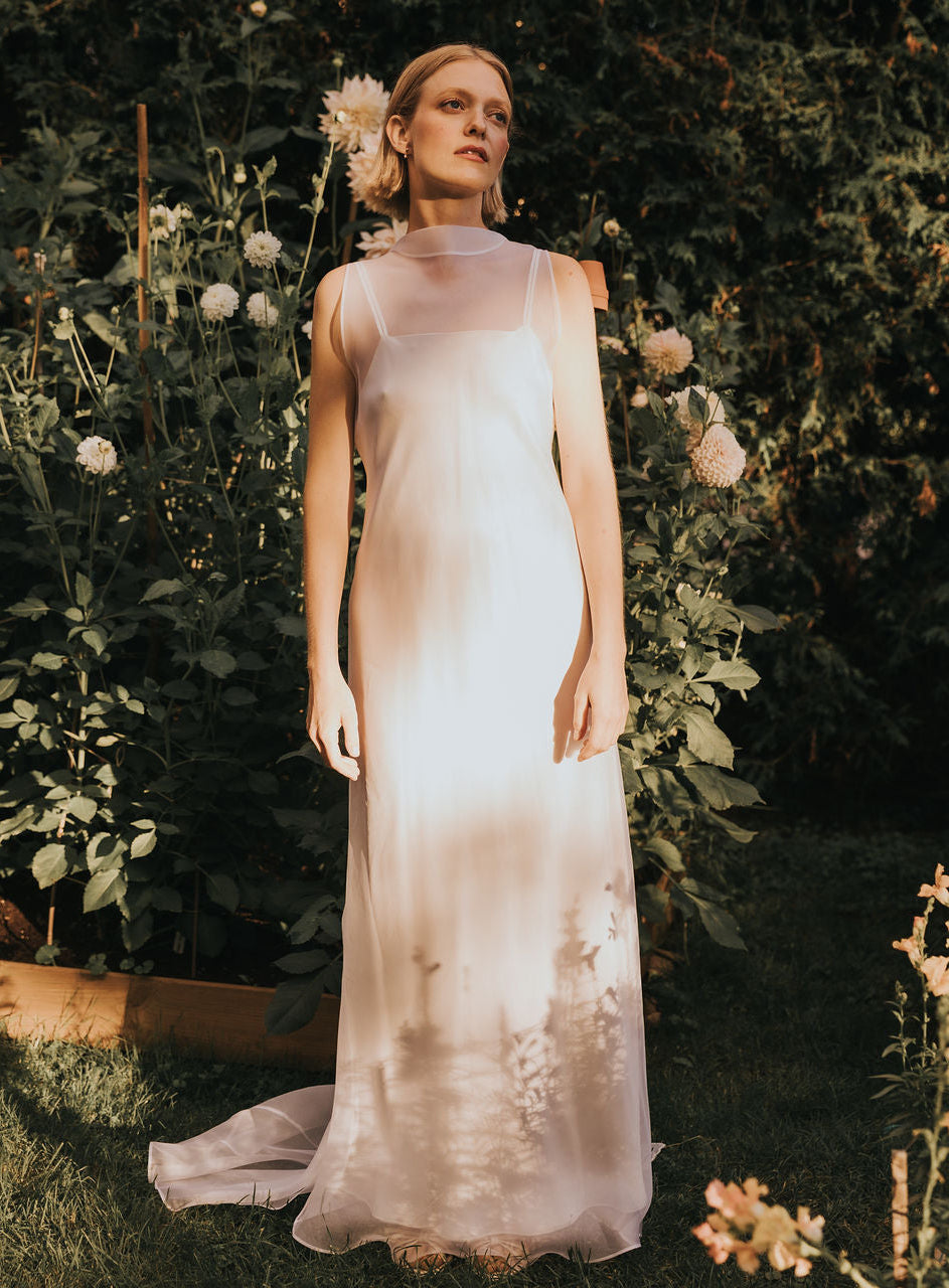 A woman stands in a garden, illuminated by sunlight and surrounded by green plants and pale flowers, wearing the Lia Dress by Noémiah—a long, sleeveless wedding gown crafted from white silk organza with a sheer neckline.