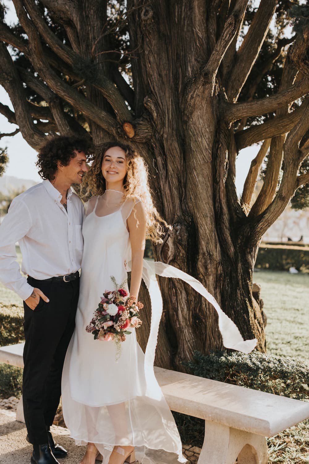 A couple stands under a large tree, smiling. The woman wears the stunning Lia Dress from Noémiah, crafted from minimalistic silk organza, as she holds a bouquet of flowers. The man complements her look with a crisp white shirt and dark pants. Nearby, a stone bench is set against the backdrop of a sunlit garden.