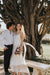 A couple stands under a large tree, smiling. The woman wears the stunning Lia Dress from Noémiah, crafted from minimalistic silk organza, as she holds a bouquet of flowers. The man complements her look with a crisp white shirt and dark pants. Nearby, a stone bench is set against the backdrop of a sunlit garden.