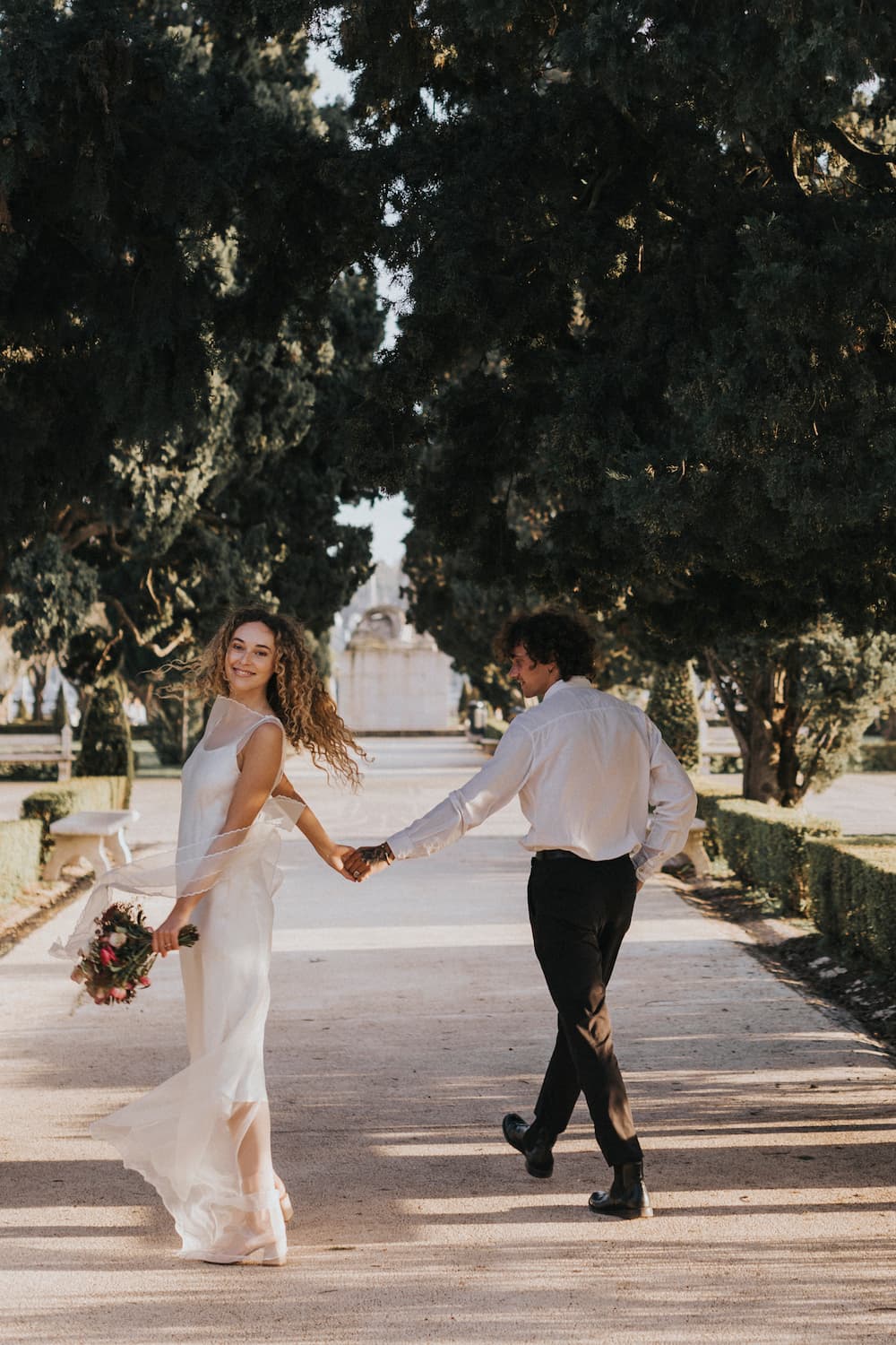 A couple strolls hand in hand along a tree-lined path in the park. The woman is elegantly dressed in the Lia Dress by Noémiah, crafted from silk organza, and she smiles over her shoulder while holding a bouquet. Her partner, wearing a white shirt and dark pants, walks behind her amidst the vibrant greenery, embodying the spirit of non-traditional brides.