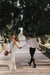 A couple strolls hand in hand along a tree-lined path in the park. The woman is elegantly dressed in the Lia Dress by Noémiah, crafted from silk organza, and she smiles over her shoulder while holding a bouquet. Her partner, wearing a white shirt and dark pants, walks behind her amidst the vibrant greenery, embodying the spirit of non-traditional brides.