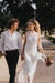 A couple walks hand in hand down a sunlit path, surrounded by trees. The woman, a non-traditional bride, wears the Lia Dress by Noémiah—an elegant piece crafted from flowing silk organza—and holds a small bouquet of flowers. The man is dressed in a white shirt and dark pants. They are smiling at each other.