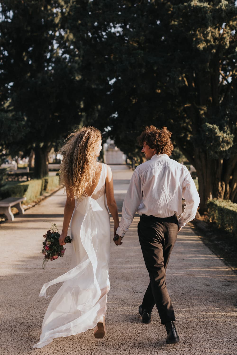 A couple walks hand in hand down a tree-lined path. The woman, exuding non-traditional bridal elegance, is adorned in the flowing Lia Dress by Noémiah and holds a bouquet of flowers, while the man complements her attire with a white shirt and dark pants. The sun casts long shadows, highlighting the serene and romantic setting.