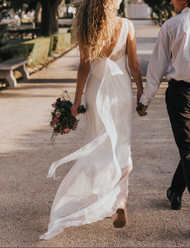 A couple walks hand in hand down a tree-lined path. The woman, exuding non-traditional bridal elegance, is adorned in the flowing Lia Dress by Noémiah and holds a bouquet of flowers, while the man complements her attire with a white shirt and dark pants. The sun casts long shadows, highlighting the serene and romantic setting.