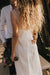 A couple stands closely in formal attire, partially visible. The woman, with long curly hair, wears the Noémiah Lia Dress made of minimalist silk organza and holds a bouquet of flowers. They are outdoors in a sunlit area with trees in the background, capturing an essence of elegance and simplicity.