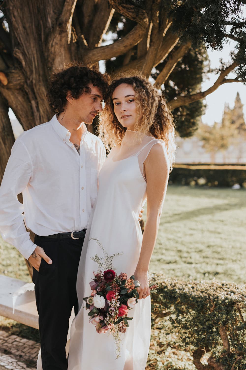 The couple stands outdoors, bathed in sunlight. The woman, radiating the allure of unconventional brides, wears the Noémiah Lia Dress made from silk organza and carries a bouquet of flowers. Beside her is a man dressed in a white shirt and dark pants. Together they are positioned before a majestic tree in the bright garden.