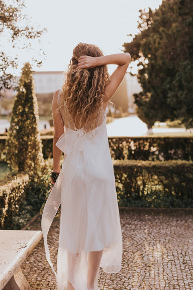 A woman with curly hair walks through a sunlit garden in the minimalist Lia Dress by Noémiah, crafted from flowing silk organza. Her back is turned as she gently touches her hair, surrounded by hedges, trees, and warm sunlight.