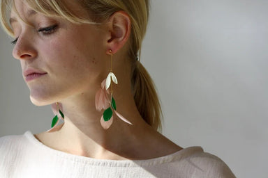 A woman with blonde hair tied back is wearing a white top. Her long, lightweight Lili Earrings by Noémiah feature pink and green leaf-like shapes. Accented by silver-plated charms, she faces to the left against a neutral background.