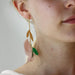 Close-up of a person wearing the Lili Earrings by Noémiah, which are lightweight and feature gold, pink, and green leaf-like elements. The minimalist design includes a gold bead and bar. The person is facing sideways, highlighting the colorful elegance inspired by high-quality feathers.