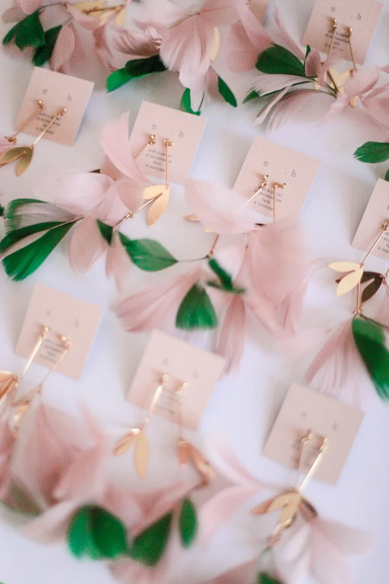 The Lili Earrings by Noémiah are presented on cards, showcasing their delicate pink feathers, green leaves, and gold accents. These lightweight earrings are artistically arranged to emphasize their elegant and whimsical design, which is complemented by silver-plated charms.