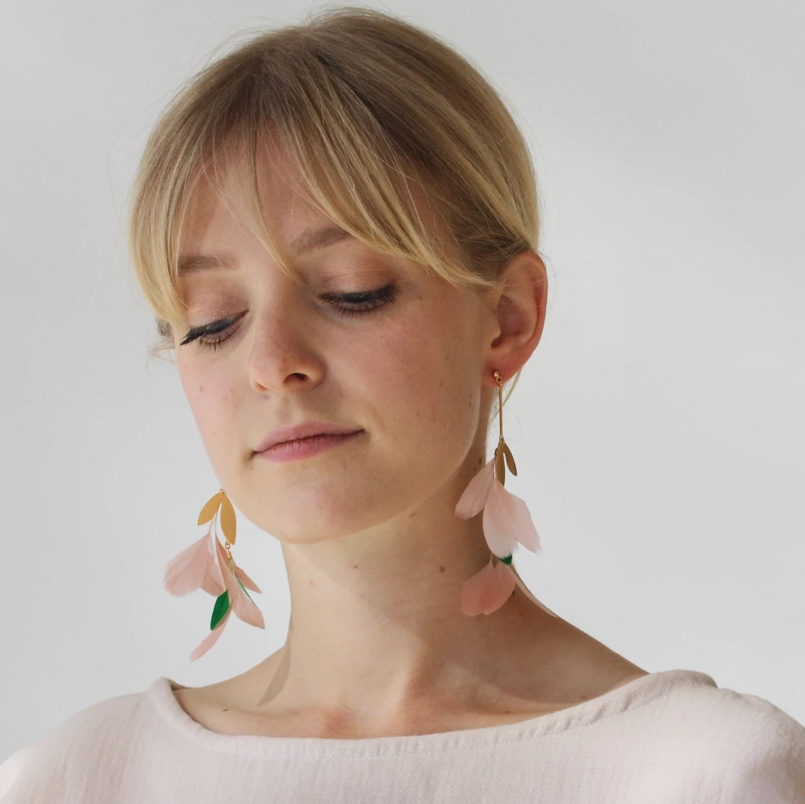 A person with light hair and a serene expression is adorned with the Lili Earrings by Noémiah, which are pink and gold and shaped like leaves. They are dressed in a light-colored top against a neutral background.