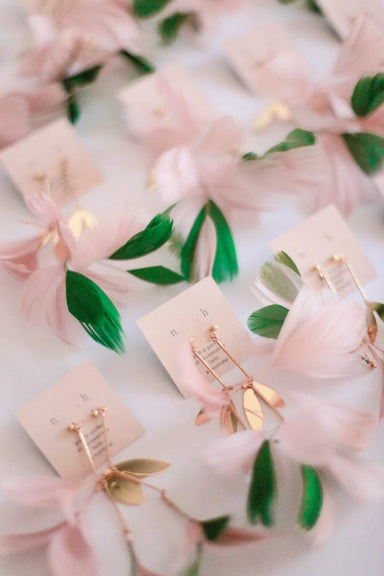 The Lili Earrings by Noémiah, featuring a delicate blend of pink and gold hues, are elegantly displayed on small cards amidst soft pink petals and green leaves. The arrangement exudes an artistic charm with its dreamy, floral theme.