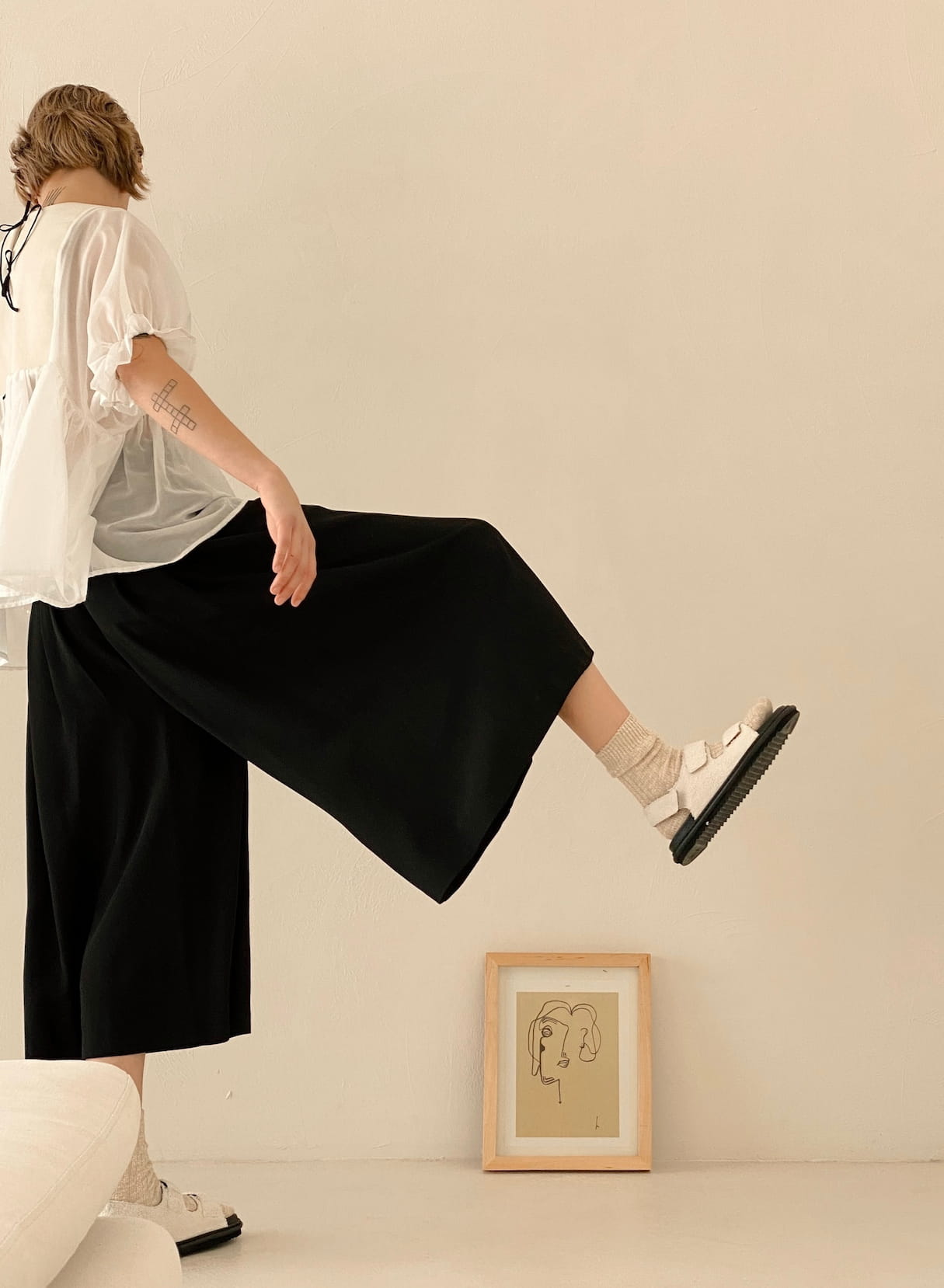 A person wearing a white blouse and Noémiah's Lison Palazzo-Pants poses with one leg raised inside a minimalistic room. They complement their outfit with white socks and sandals, while a line drawing artwork leans against the wall, creating a simple and neutral setting.