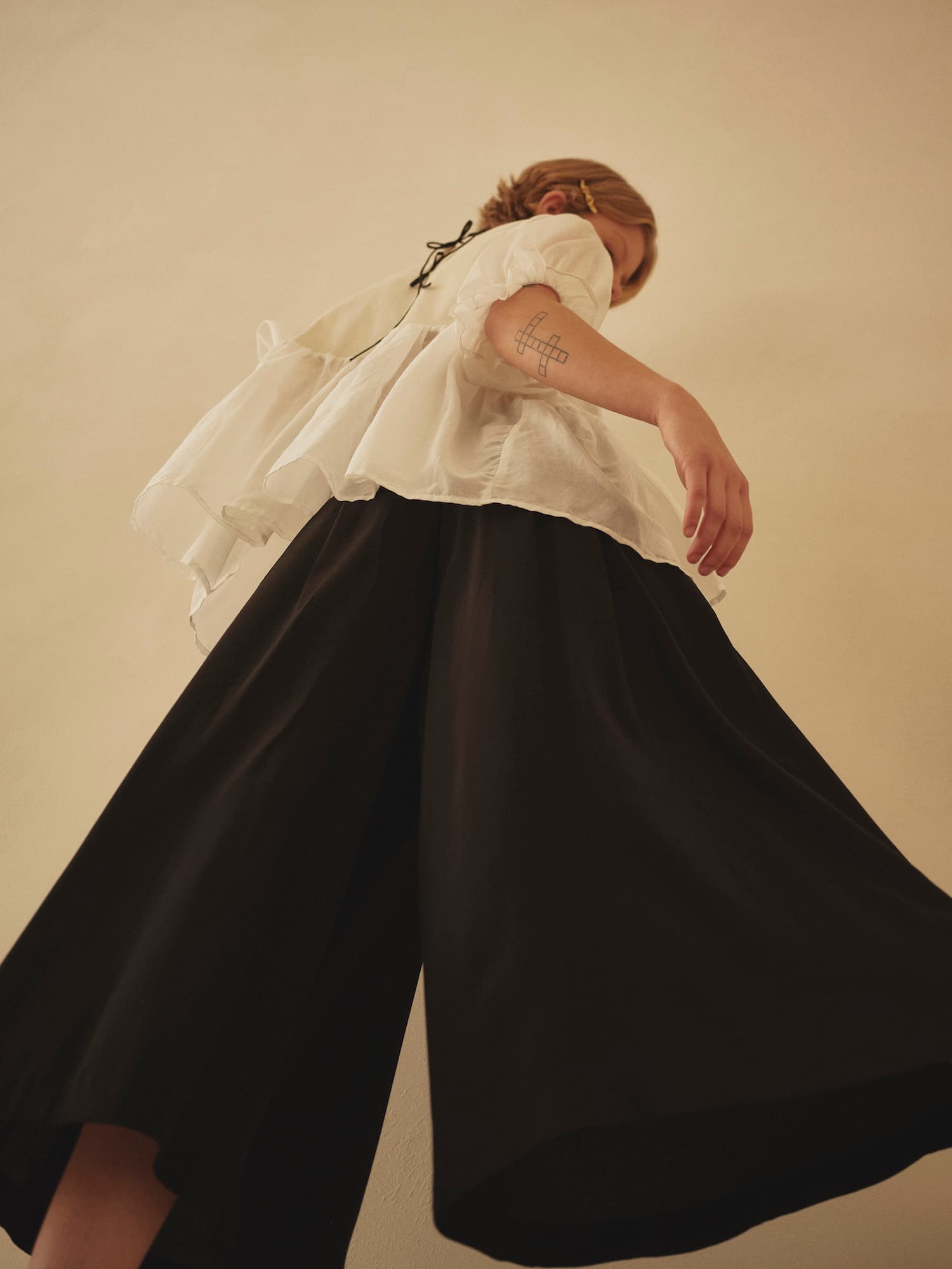 A person in a flowing white top and Noémiah's Lison Palazzo-Pants stands with an arm outstretched. The camera angle is low, capturing the outfit's graceful movement. They have a tattoo on their arm and light brown hair tied back, set against a plain, warm-toned wall.