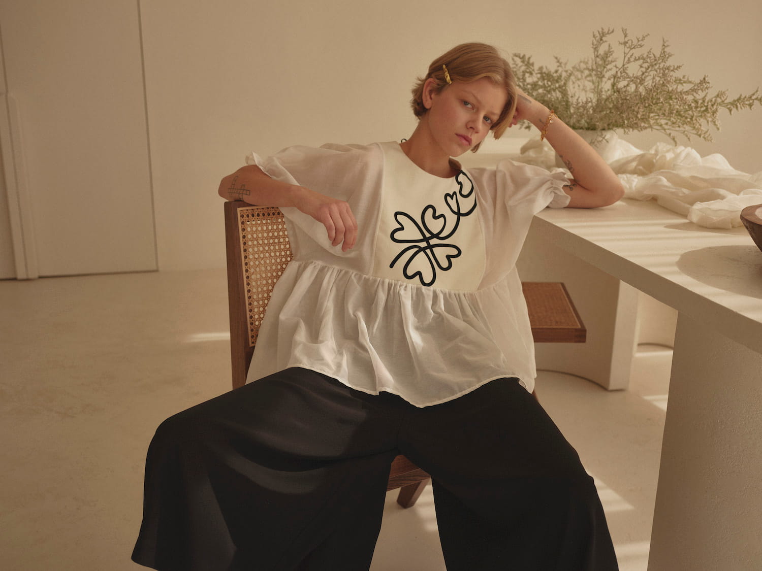 In a minimalist room, an individual with short hair is seated, adorned in a white top featuring black abstract designs and Noémiah's Lison Palazzo-Pants crafted from silk crepe. They rest one elbow on a table, wearing a relaxed, contemplative expression.