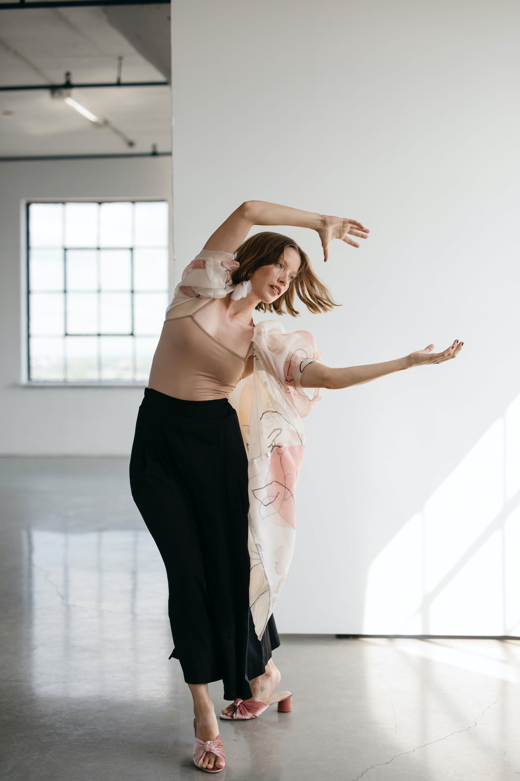 A woman dances gracefully in a sunlit room with concrete floors. She wears a beige top, a flowing silk crepe shawl, Lison Palazzo-Pants by Noémiah, and pink sandals. Her arms are extended and her hair moves as she poses expressively.