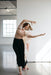 A woman dances gracefully in a sunlit room with concrete floors. She wears a beige top, a flowing silk crepe shawl, Lison Palazzo-Pants by Noémiah, and pink sandals. Her arms are extended and her hair moves as she poses expressively.