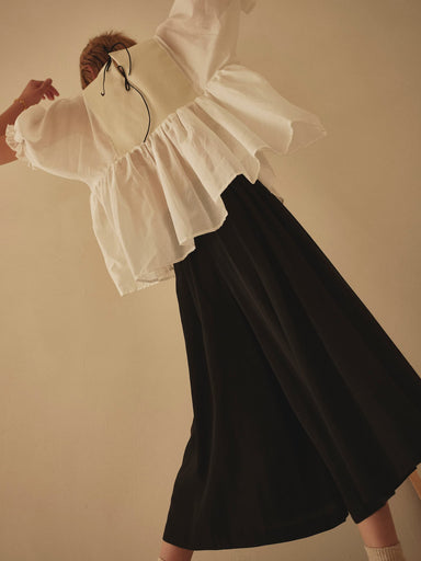 A person wearing a white, ruffled blouse and Lison Palazzo-Pants by Noémiah is dancing energetically with arms raised. The angle is from below, providing a dynamic and playful perspective against a plain, muted wall backdrop.