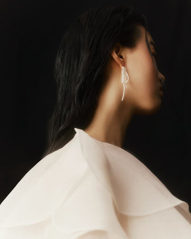 A woman with long dark hair wears a white, ruffled top, gazing to the right against a dark background. She is adorned with Noémiah's Lluvia Earrings, which feature delicate freshwater pearls. The image exudes a soft focus and elegant, minimalistic style, reminiscent of NINAJANVIER's signature approach.