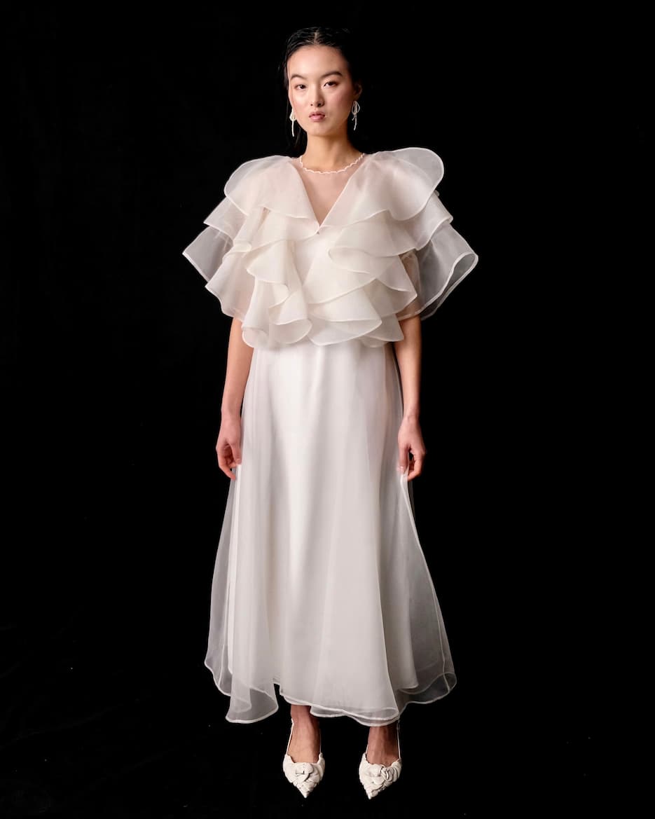 A woman stands against a black background wearing a flowing white dress with layered, ruffled sleeves. Adorning her ears are the Lluvia Earrings by Noémiah, complementing her white pointed shoes, while her hair is elegantly pulled back. This ensemble reflects the grace and sophistication of NINAJANVIER.