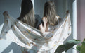 Two women with long hair stand side by side, facing away from the camera, holding a light silk charmeuse scarf featuring the floral pattern of the Long Swimmers Scarf by Noémiah. Sunlight filters through a window, casting gentle shadows on the wall, while Paule T.B.'s plant leaves can be seen in the foreground.