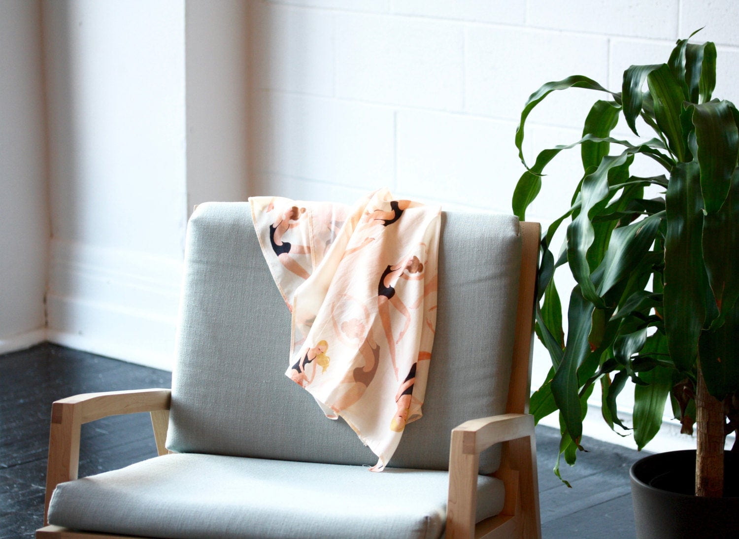 A Long Swimmers Scarf from Noémiah, adorned with abstract patterns, drapes over a minimalist wooden chair that features a light gray cushion. Beside the chair sits a leafy green plant against a white brick wall backdrop, capturing the elegance of Paule T.B. design.