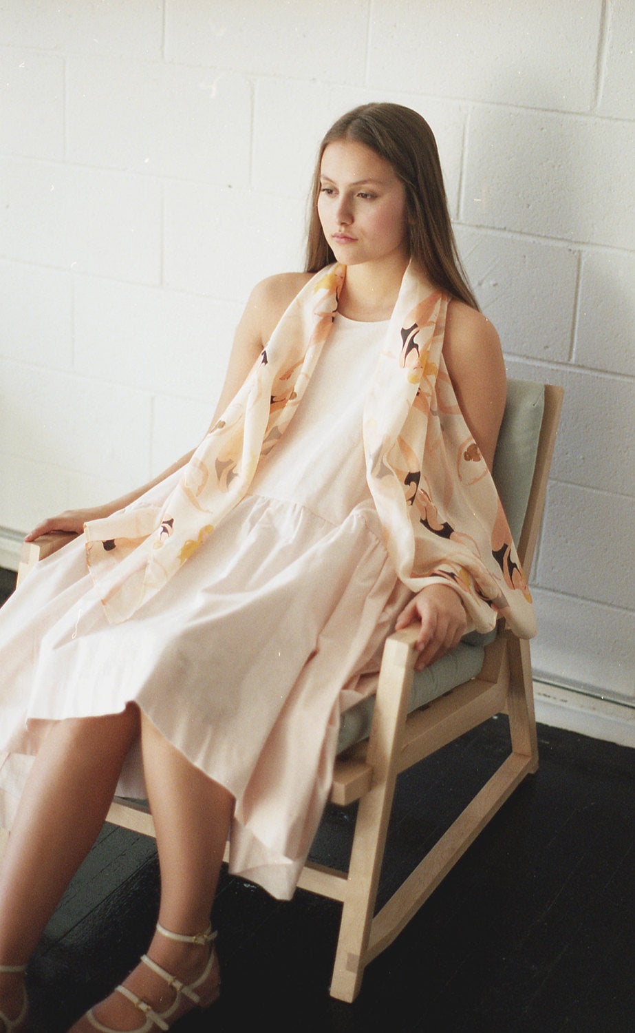 A young woman with long brown hair sits in a light wooden chair, wearing a light pink dress complemented by a flowy, patterned Long Swimmers Scarf from Noémiah. The setting is a bright room with a white brick wall, where her calm and contemplative expression beautifully complements the elegance of her silk shawl.