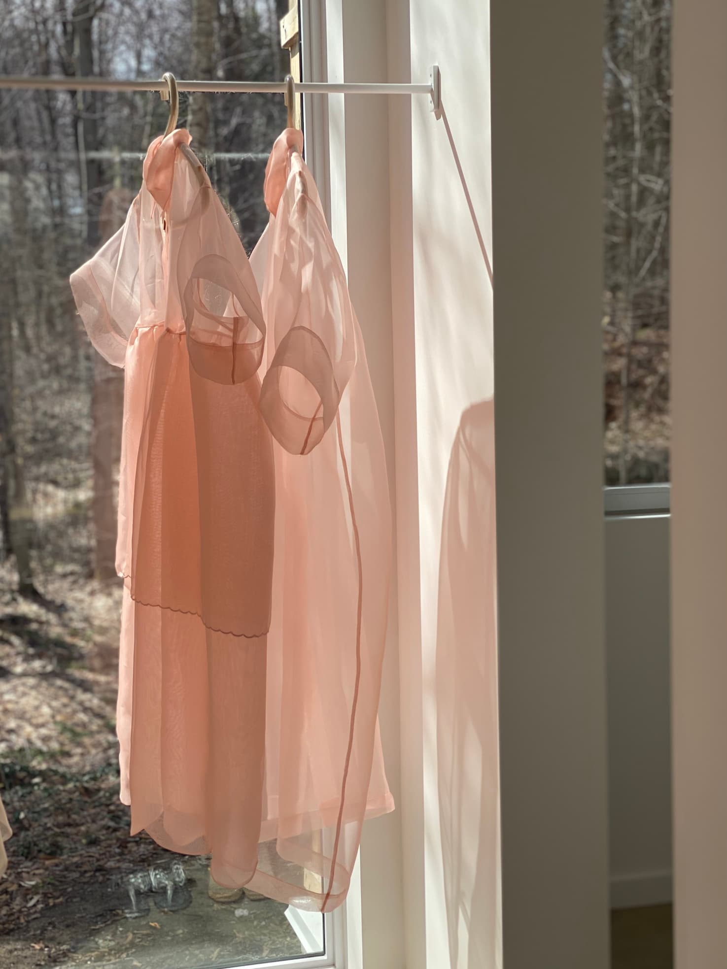 Two Lou Shirt Dresses, designed by Noémiah and crafted from sheer pink silk organza, hang on a slender bar in a sunlit room. They sway gently in the breeze, casting soft shadows on the wall. Outside, barren trees stand against the winter scene visible through the window.