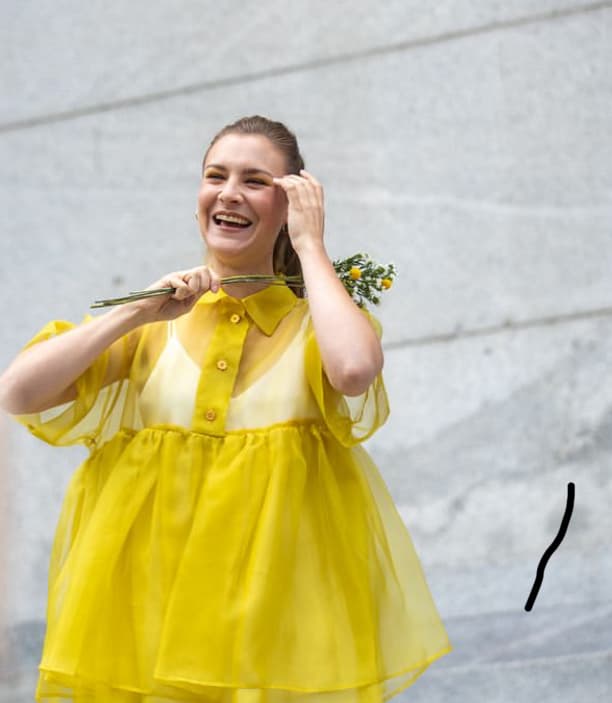 A woman stands against a light gray wall, smiling and holding a small bouquet of yellow flowers. She is wearing the Lou Shirt Dress from Noémiah, a bright yellow, relaxed fit dress crafted from delicate silk organza.