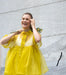 A woman stands against a light gray wall, smiling and holding a small bouquet of yellow flowers. She is wearing the Lou Shirt Dress from Noémiah, a bright yellow, relaxed fit dress crafted from delicate silk organza.