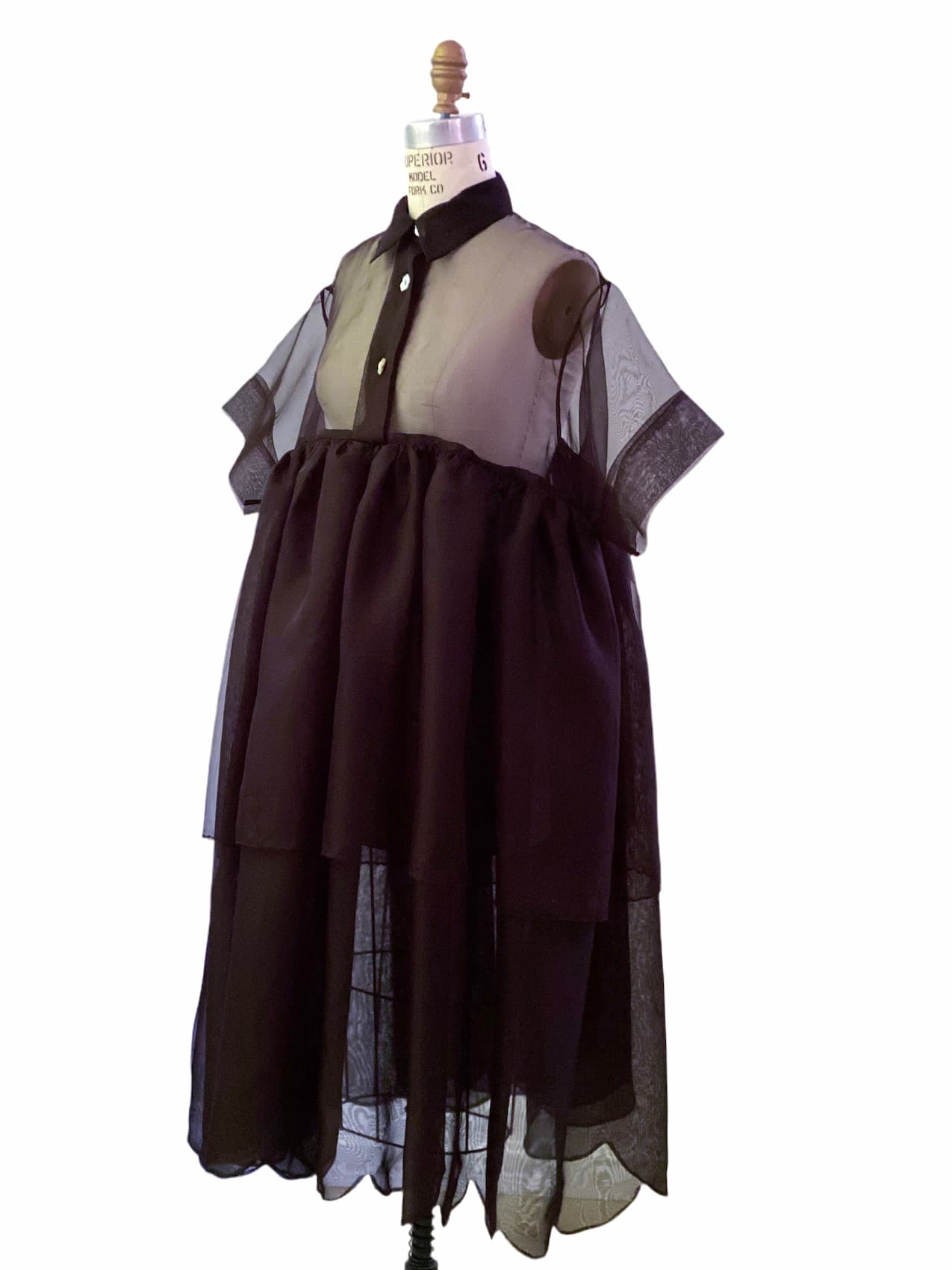 The Lou Shirt Dress by Noémiah is elegantly showcased on a dress form, featuring a sheer black design with short sleeves and layered tulle. Crafted from silk organza, this garment includes a collar, button-up top, and voluminous skirt, highlighting a captivating blend of textures and transparency.