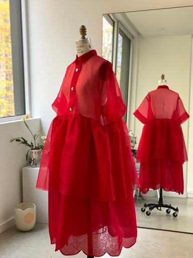 The Lou Shirt Dress by Noémiah, a striking creation with its red tulle and sheer top layered over a flowing skirt, is elegantly showcased on a mannequin. Its beauty is mirrored in a large glass nearby, set against the room's white walls and the colorful autumn leaves visible through the window. The room is also adorned with a white vase and a small decorative plant, contributing to an atmosphere of relaxed fit luxury.