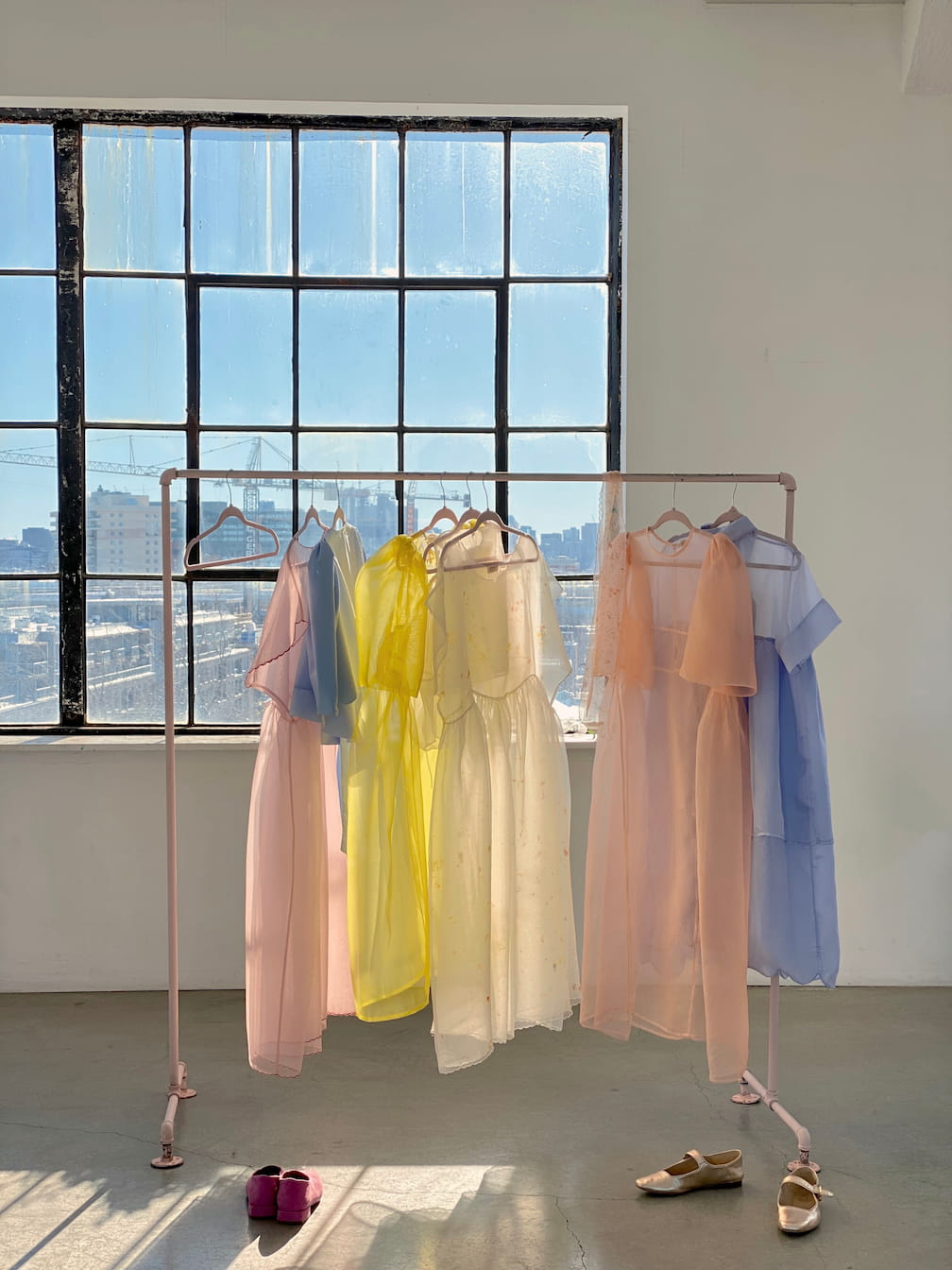 A clothing rack showcases pastel-colored dresses in pink, yellow, and blue, featuring a relaxed fit Lou Shirt Dress made of silk organza by Noémiah, in front of a large sunlit industrial window. Below the rack is a pair of purple slippers and beige sandals resting on the gray floor.