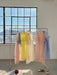 A clothing rack showcases pastel-colored dresses in pink, yellow, and blue, featuring a relaxed fit Lou Shirt Dress made of silk organza by Noémiah, in front of a large sunlit industrial window. Below the rack is a pair of purple slippers and beige sandals resting on the gray floor.