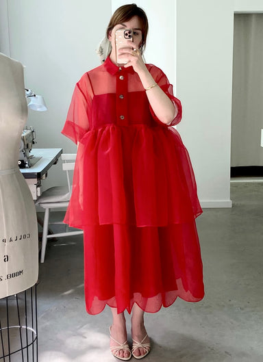 A person takes an indoor mirror selfie wearing the Lou Shirt Dress by Noémiah in a relaxed red silk organza style, paired with light sandals and gold rings. A white mannequin and chair can be seen in the background.