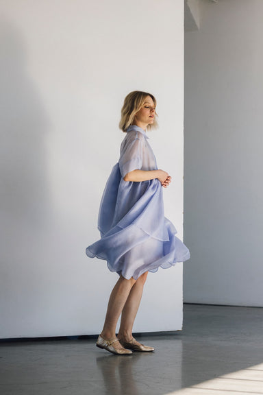 A woman in the Lou Shirt Dress by Noémiah, a sheer, oversized shirt dress in light blue, stands elegantly barefoot in a minimalist room with white walls and concrete flooring. Gazing off to the side, she embodies an air of elegance and simplicity as soft shadows fall on the floor.