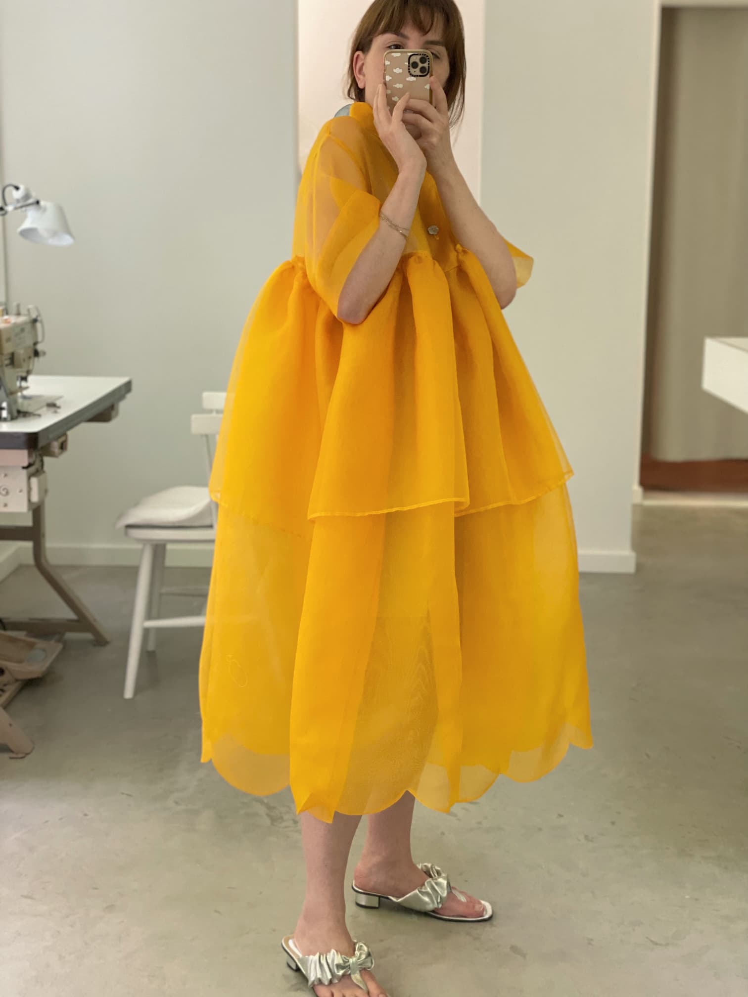 In a room filled with sewing equipment and neutral-toned floors and walls, a person takes a mirror selfie while wearing the Noémiah Lou Shirt Dress. The flowing, layered silk organza design in a vibrant yellow hue pairs perfectly with their slip-on shoes as they hold their phone.