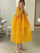 In a room filled with sewing equipment and neutral-toned floors and walls, a person takes a mirror selfie while wearing the Noémiah Lou Shirt Dress. The flowing, layered silk organza design in a vibrant yellow hue pairs perfectly with their slip-on shoes as they hold their phone.