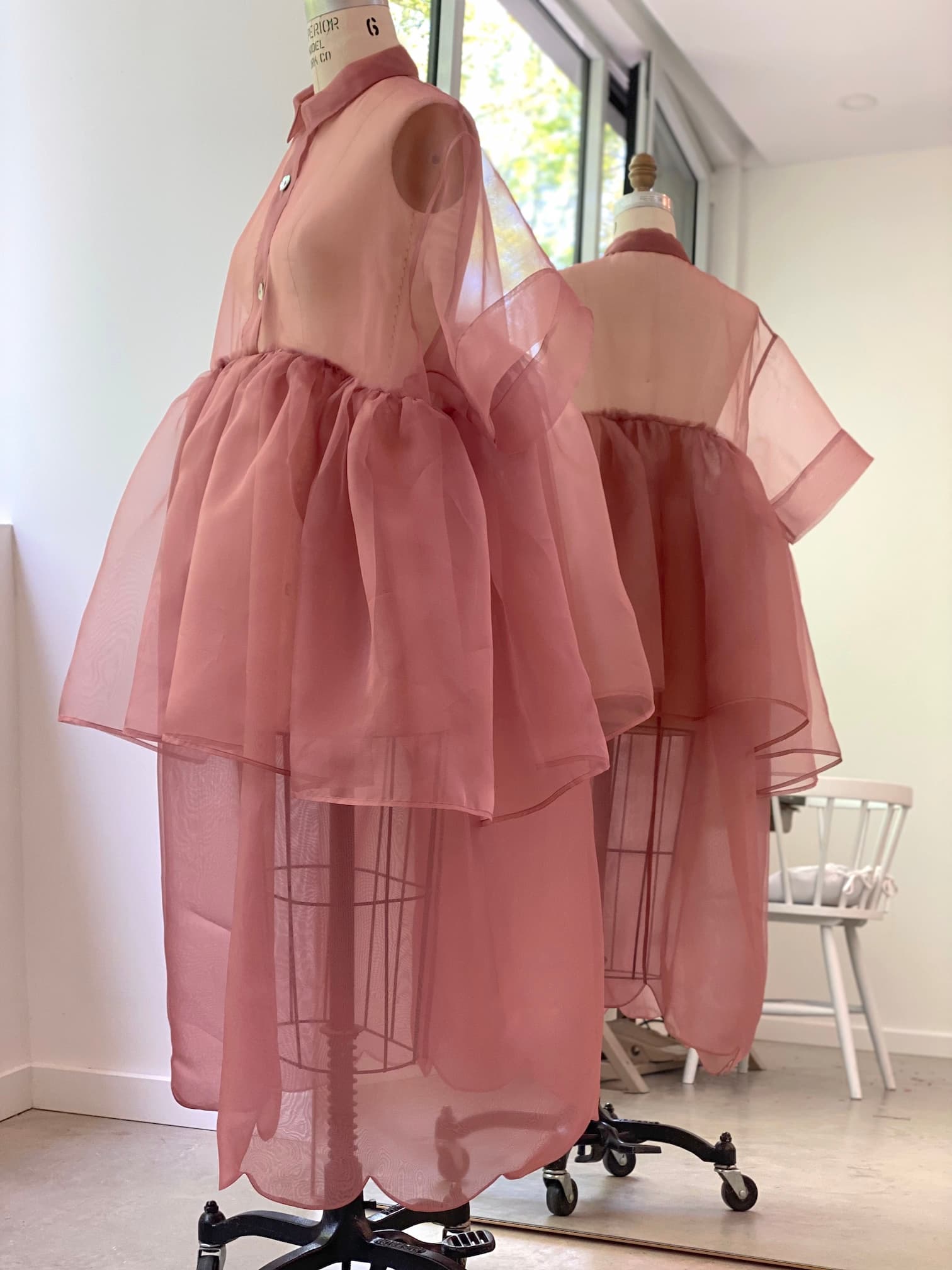 A dress form showcases the Lou Shirt Dress by Noémiah, featuring a sheer pink silk organza design with a voluminous, layered skirt. The sleeveless, button-up top resembles an oversized shirt dress. Positioned before a large window with white walls, the ensemble is elegantly reflected in a nearby mirror.