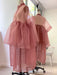 A dress form showcases the Lou Shirt Dress by Noémiah, featuring a sheer pink silk organza design with a voluminous, layered skirt. The sleeveless, button-up top resembles an oversized shirt dress. Positioned before a large window with white walls, the ensemble is elegantly reflected in a nearby mirror.