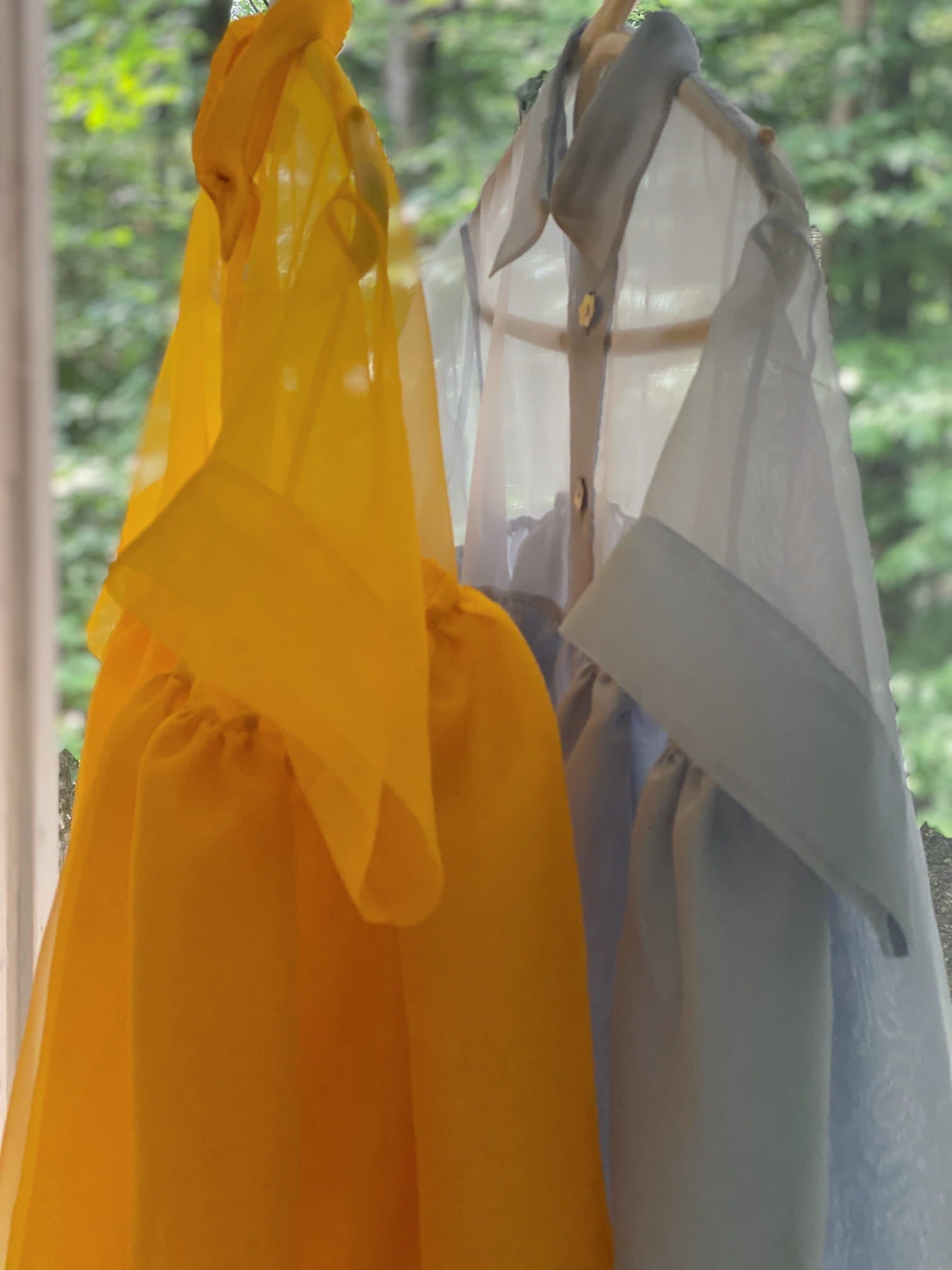 Two Lou Shirt Dresses from Noémiah, made of silk organza, hang side by side on hangers. One is a vibrant yellow and the other a soft blue, both featuring long, flowing sleeves and a relaxed fit. They are set against a blurred background of green foliage visible through a window.