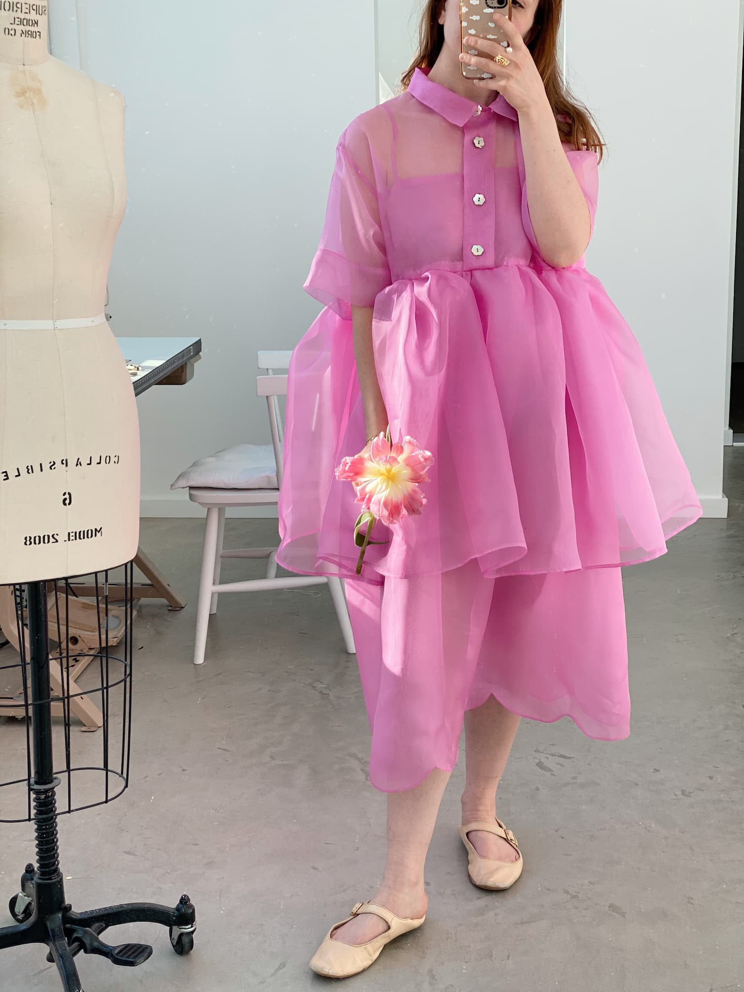 A person poses gracefully with a dress form while wearing the Lou Shirt Dress in pink sheer silk organza by Noémiah. Holding a flower, they snap a selfie in the room, which features a stool and a neutral-toned floor, capturing the essence of relaxed fit style.